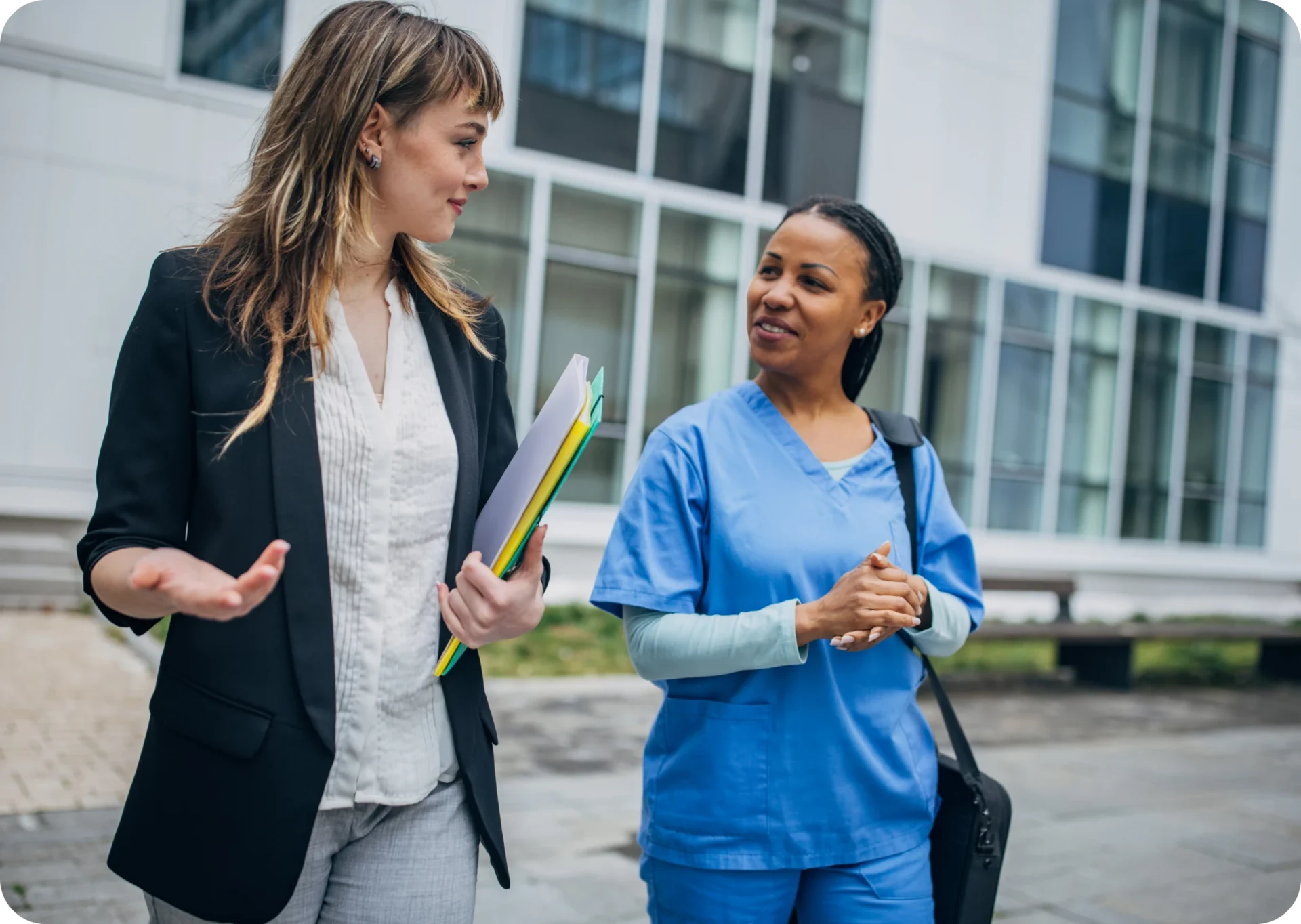 Two women walking and conversing outside a building; one is in a black blazer holding folders, the other in blue scrubs with a bag.